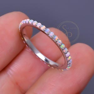 Op de afbeelding: Een dunne zilverkleurige metalen ring met een doorlopende rij kleine, ronde, iriserende witte opalen. De stenen vertonen subtiele kleurflitsen en de ring heeft een gepolijste afwerking. Een cirkelvormig logo met de letters SP is zichtbaar.