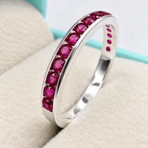 Peut inclure: Une bague en métal argenté dotée d'un sertissage rail avec une rangée de pierres précieuses rondes d'un rose magenta vif. L'anneau métallique présente une finition polie et réfléchissante, et les pierres sont alignées uniformément.