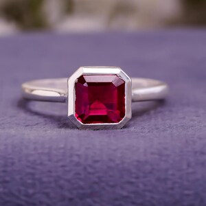 May include: A silver ring with a large, rectangular, deep red gemstone. The gemstone is set in a bezel setting, and the band is smooth and polished. The ring is displayed on a purple surface.