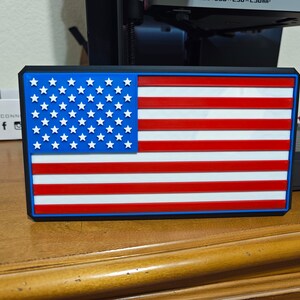 以下が含まれることがあります： 青い縁取りのある3Dプリントされたアメリカ国旗のパッチ。旗は赤、白、青のプラスチック製で、表面にテクスチャがあります。