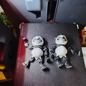 Peut inclure: Deux figurines de panda articulées imprimées en 3D avec une fourrure noire et blanche, allongées sur une surface noire texturée. Les pandas ont des yeux bleus et verts et sont posés avec leurs bras et leurs jambes tendus.