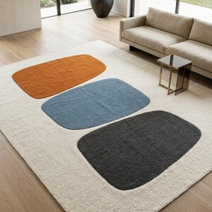 May include: A rectangular area rug with a neutral beige background and three large, rounded rectangular shapes in burnt orange, blue, and charcoal gray. The rug is placed in a modern living room setting.