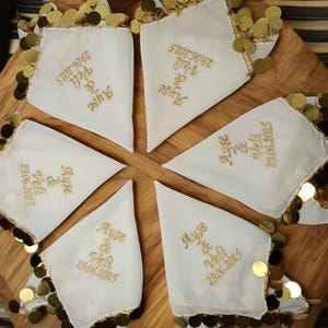 Puede incluir: Seis pañuelos blancos con ribete de lentejuelas doradas están dispuestos sobre una superficie de madera. Cada pañuelo tiene texto bordado en oro que dice "Ayse & Veli 29.06.2025". Los pañuelos están dispuestos en círculo.