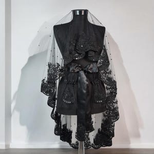 May include: Black lace veil with beaded embellishments. The veil is draped over a dress form and has a satin ribbon tied around the waist.