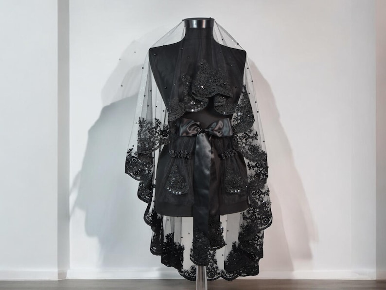 May include: Black lace veil with beaded embellishments. The veil is draped over a dress form and has a satin ribbon tied around the waist.