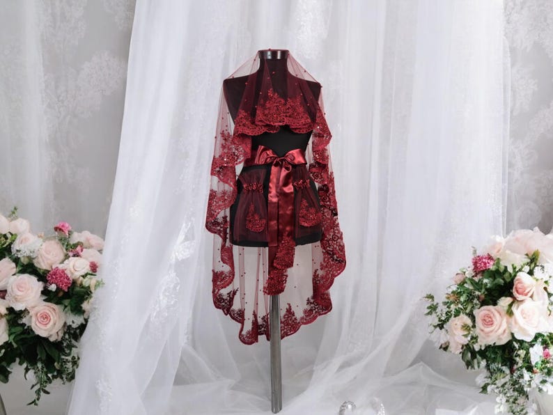 May include: A burgundy lace bridal veil with a satin ribbon and beaded embellishments. The veil is displayed on a mannequin against a white backdrop.