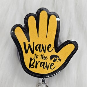 May include: A yellow hand-shaped badge reel with the text "Wave to the Brave" in black. The badge reel has a black border and a clear plastic clip and button. The background is a fluffy white surface.