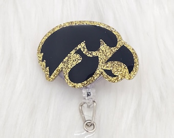 May include: A badge reel featuring a black and gold glitter eagle head logo. The design is encased in a smooth, glossy finish with a gold glitter border. It includes a clear plastic retractable cord and a metal clip for attaching identification cards.