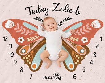 May include: A light pink milestone blanket featuring a large, colorful butterfly graphic with orange, teal, and white floral patterns. The text reads Today Zelie Is and months, with numbers 1 through 12 arranged around the butterfly wings.