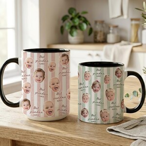 May include: Two ceramic mugs with black handles and rims. The mugs are decorated with baby faces and the text "Best grandma ever" on a striped background. One mug is larger than the other.