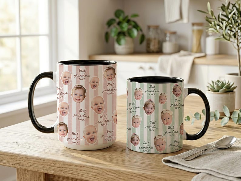 May include: Two ceramic mugs with black handles and rims. The mugs are decorated with baby faces and the text "Best grandma ever" on a striped background. One mug is larger than the other.