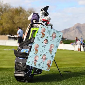 May include: A light blue golf towel featuring cartoon illustrations of children playing golf, with the text "Best Dad Ever" repeated throughout. The towel is displayed next to a golf bag on a green golf course.