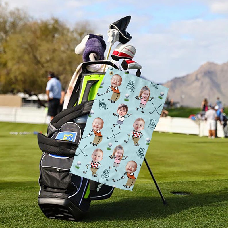 May include: A light blue golf towel featuring cartoon illustrations of children playing golf, with the text "Best Dad Ever" repeated throughout. The towel is displayed next to a golf bag on a green golf course.