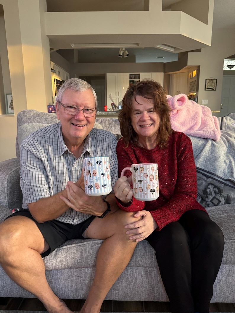 May include: Two people are seated on a gray sofa, each holding a white mug. The mugs feature a pattern of baby faces and bows. One mug has a pink handle, the other has a blue handle. The person on the left is wearing a plaid shirt and black shorts.