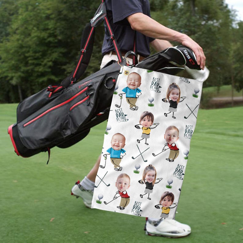 May include: A golf bag with a black and red design is carried on a person's shoulder. A white golf towel with cartoon images of children and the text "BEST DAD BY FAR" is hanging from the bag. The scene takes place on a green golf course.