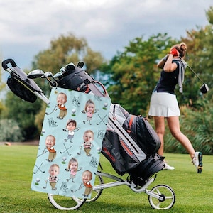 May include: A golf bag on a cart with a personalized towel featuring cartoon babies and the text "Best Dad Ever." A woman swings a golf club in the background on a green golf course.