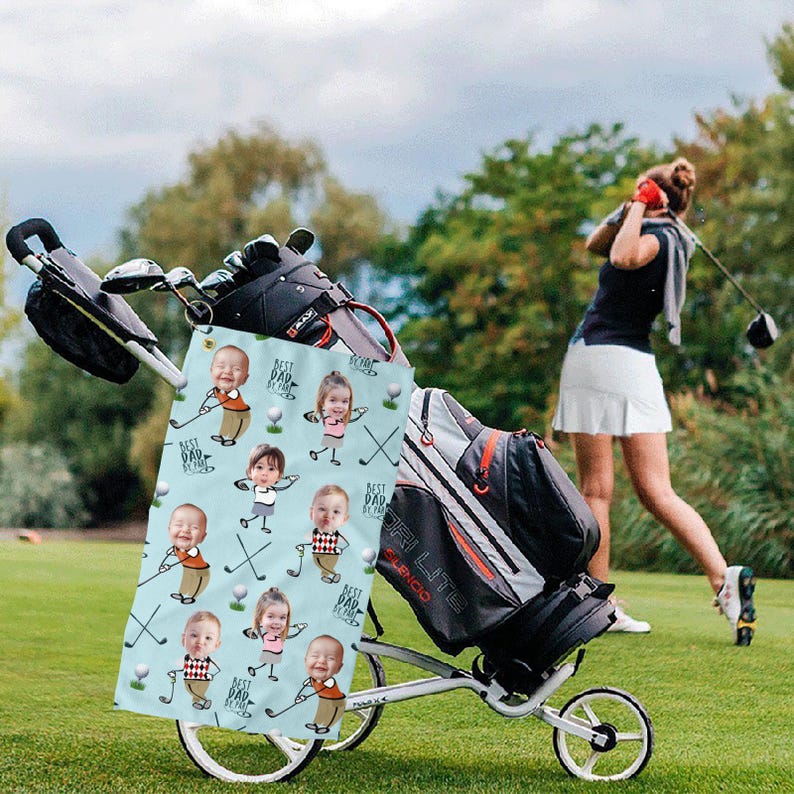 May include: A golf bag on a cart with a personalized towel featuring cartoon babies and the text "Best Dad Ever." A woman swings a golf club in the background on a green golf course.