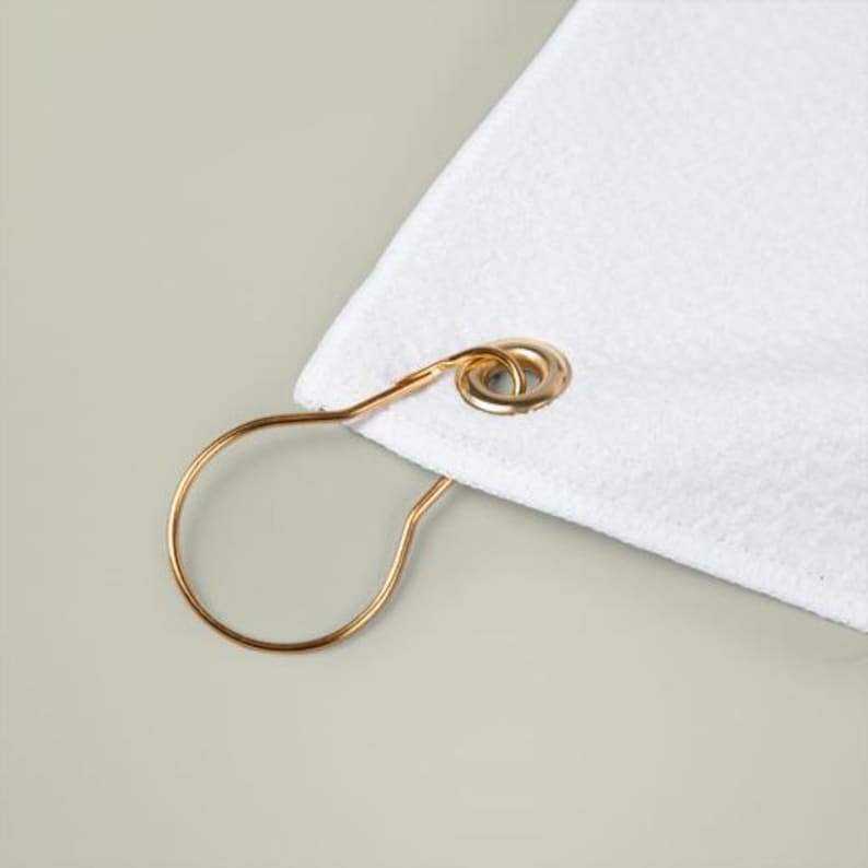May include: Close-up of a white, textured towel corner with a gold-colored metal shower curtain ring attached. The ring has a circular design and is attached to the towel with a gold-colored grommet.