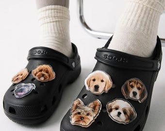 May include: A pair of black clogs featuring multiple dog-themed charms attached to the top surface. The charms are shaped like various dog breeds, including a golden retriever, a beagle, a shih tzu, a yorkshire terrier, and other canine portraits.