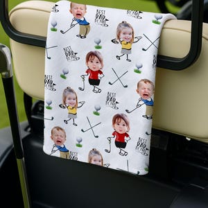 May include: White golf towel featuring a repeating pattern of children with golf clubs, golf balls, and crossed golf clubs. The text "BEST DAD BY PAR" is also printed on the towel. The towel is draped over a golf cart.