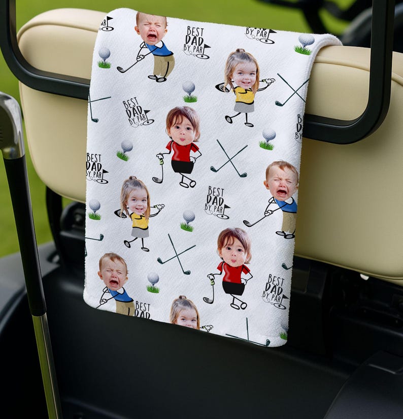 May include: White golf towel featuring a repeating pattern of children with golf clubs, golf balls, and crossed golf clubs. The text "BEST DAD BY PAR" is also printed on the towel. The towel is draped over a golf cart.