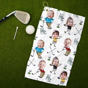 May include: White golf towel featuring a repeating pattern of cartoon children with golf clubs and golf balls. The towel has the text "BEST DAD BY PAR" in a circular design. A golf club, tee, and ball are on a green surface.