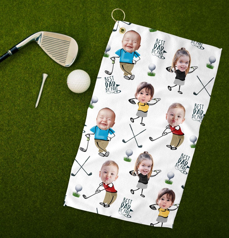 May include: White golf towel featuring a repeating pattern of cartoon children with golf clubs and golf balls. The towel has the text "BEST DAD BY PAR" in a circular design. A golf club, tee, and ball are on a green surface.