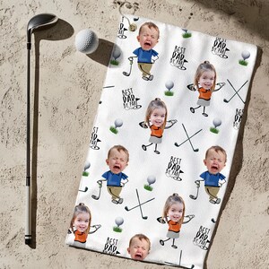 May include: White golf towel featuring a repeating pattern of cartoon babies and golf-related graphics. The towel has the text "BEST DAD BY PAR" and golf clubs. A golf club and ball are next to the towel.