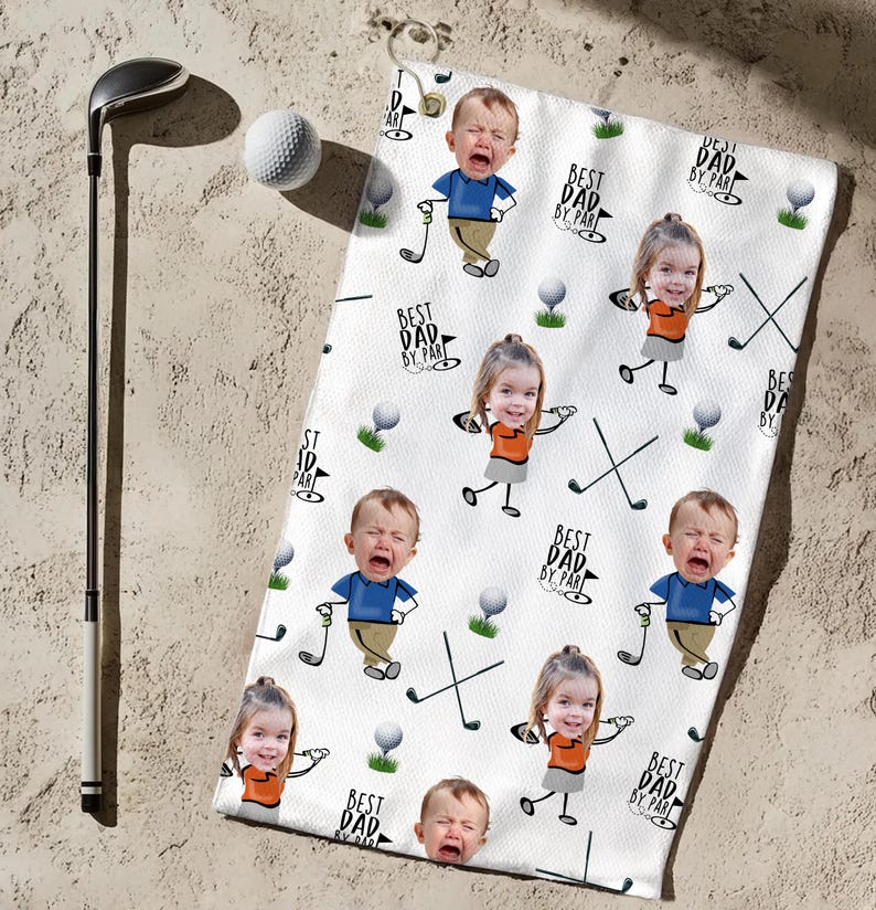 May include: White golf towel featuring a repeating pattern of cartoon babies and golf-related graphics. The towel has the text "BEST DAD BY PAR" and golf clubs. A golf club and ball are next to the towel.