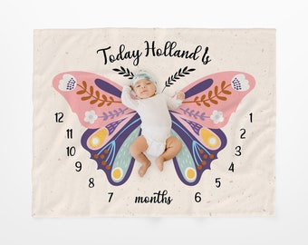 May include: A baby girl lies on a cream-colored blanket with a butterfly design. The butterfly has pink, purple, blue, and yellow wings with floral accents. The blanket has numbers 1-12 on one side and the words "Today Holland is" and "7 months" on the other side.