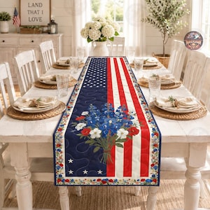 May include: A quilted table runner featuring a patriotic American flag design with red, white, and blue stripes and stars. The centre displays a bouquet of blue, white, and red flowers, bordered by a floral pattern on a white background.
