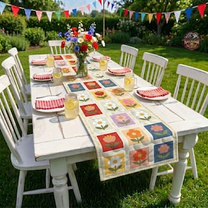 May include: A rectangular table runner featuring a grid of colourful floral squares in shades of red, orange, yellow, pink, and blue. The runner is placed on a white wooden table with matching chairs, glass jars, and red chequered napkins.