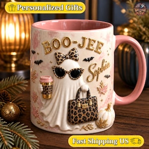 May include: A ceramic mug with a pink interior and handle, featuring a raised 3D design of a ghost wearing leopard print sunglasses, a matching bow, and holding a leopard print coffee cup and handbag. The text reads BOO-JEE and Sophia.