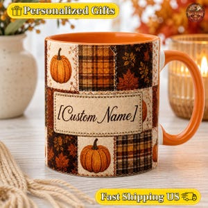 Peut inclure: Un mug en céramique avec un intérieur et une anse orange, orné d'un motif patchwork de citrouilles, de feuilles d'automne et de carreaux dans des tons marron, orange et crème. Un écusson central affiche le texte Custom Name en noir.
