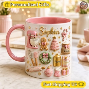 Op de afbeelding: Een witte keramische mok met een roze binnenkant en handvat, voorzien van de naam Sophia in gouden letters. De buitenkant is versierd met reliëf-illustraties van een roze mixer, peperkoekmannetje, kerstboom, meelzak, krans, deegroller, eierpunch-pak en een cadeau.