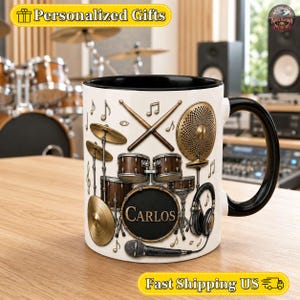 May include: A white ceramic mug with a black handle and rim, featuring a graphic print of a drum kit, drumsticks, a microphone, and headphones. The centre of the bass drum is circular and displays the name Carlos in gold lettering.