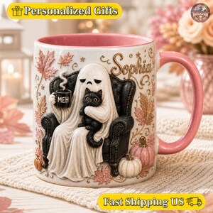 Könnte beinhalten: Ein Keramikbecher mit rosa Innenseite und Henkel, der einen Geist in einem schwarzen Sessel zeigt, der eine schwarze Katze und eine kleine Tasse mit der Aufschrift MEH hält. Das Design umfasst den Namen Sophia, Herbstblätter und Kürbisse.