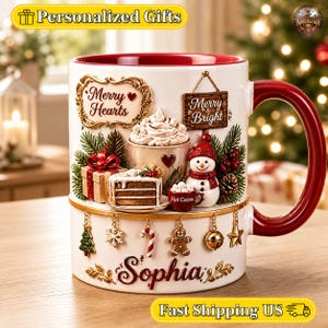 Könnte beinhalten: Ein weißer Keramikbecher mit rotem Innenraum und Henkel, verziert mit einer weihnachtlichen 3D-Szene aus Schneemann, Kuchenstück, heißer Schokolade und Geschenk. Der Becher trägt die Aufschriften Merry Hearts, Merry & Bright sowie den Namen Sophia.