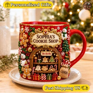 May include: A red ceramic mug featuring a three-dimensional holiday scene with a gingerbread shop, snowmen, and Christmas trees. The text "SOPHIA'S COOKIE SHOP" is centred in a gold frame above a shop window display with "HO HO HO" signage.