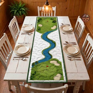 May include: A rectangular table runner featuring a printed golf course design with green fairways, sand bunkers, and a winding blue river. The fabric has a quilted texture and is centered on a white wooden dining table with place settings.