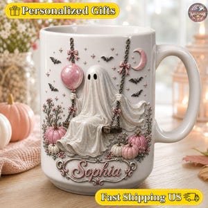 Könnte beinhalten: Ein weißer Keramikbecher mit einer erhabenen Illustration eines Geistes auf einer Schaukel, der einen rosa Ballon hält. Das Design zeigt rosa Kürbisse, kleine Fledermäuse, eine Mondsichel und den Namen Sophia in eleganter Schrift unten am Becher.