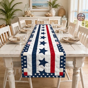 Op de afbeelding: Een doorgestikte tafelloper met een patriottisch ontwerp met verticale strepen in rood, wit en blauw, geaccentueerd door een herhalend patroon van marineblauwe sterren op een witte achtergrond, afgewerkt met een rand van kleine witte sterren.
