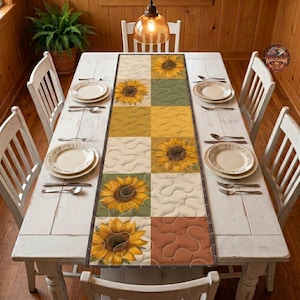 May include: A rectangular quilted table runner featuring a grid pattern of sunflowers and solid blocks in shades of yellow, green, and brown. The runner is centred on a white wooden dining table with four white chairs and two white plates.