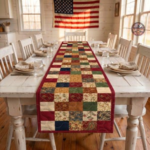 May include: A rectangular quilted table runner featuring a patchwork design of squares in shades of red, green, blue, cream, and brown with various floral and paisley patterns, finished with a solid dark red border and decorative stitching.