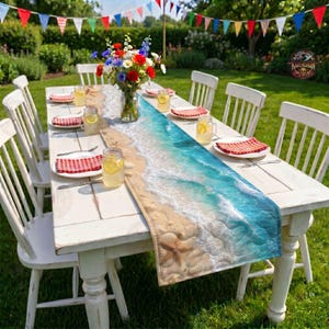 May include: A rectangular table runner featuring a printed beach scene with sand, a starfish, and turquoise ocean waves. The runner is placed on a white wooden table with white chairs, red and white checkered napkins, and a glass vase of flowers.