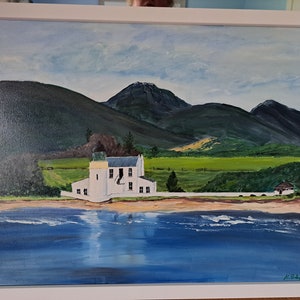 Puede incluir: Una pintura de una casa blanca con techo azul, situada en un fondo de colinas verdes y una masa de agua azul. El cielo es azul claro con nubes blancas. La pintura está enmarcada en blanco.