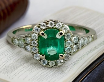 Peut inclure: Une bague en or avec une grande émeraude entourée de diamants plus petits. La bague est posée sur un livre blanc.