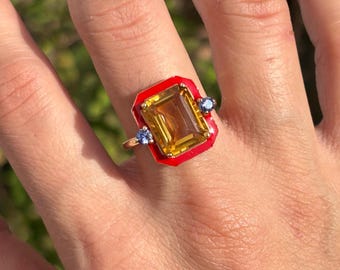 Op de afbeelding: Een gouden ring met een grote, rechthoekige gele edelsteen, gezet in een rood emaille frame. Twee kleine blauwe edelstenen flankeren de hoofdsteen. De ring wordt getoond op een hand met roze nagellak.