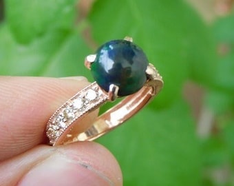Op de afbeelding: Roségouden ring met een grote, donkerblauwe edelsteen en kleine, heldere accentstenen. De band heeft een rij kleine, heldere stenen. De ring is geplaatst tegen een vage groene achtergrond.