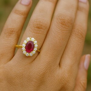 Op de afbeelding: Een gouden ring met een ovale robijn als middensteen, omringd door een halo van kleine diamanten. De ring heeft een fijne gouden band, met een klassiek en elegant ontwerp. De ring wordt getoond op de hand van een persoon.